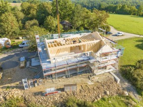 Timber frame construction with scaffolding and crane, embedded in a wooded landscape, timber house