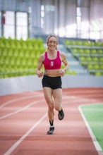 Female athlete smiles while running on a red track in a hall, middle distance runner Katharina