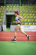 A motivated athlete runs past green stands on an indoor track, middle distance runner Katharina