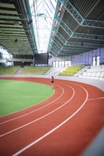 A person runs on an oval, red-green running track of a spacious indoor sports hall, middle distance
