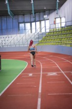 A runner with pink hair jogs on a red track in an indoor stadium, middle distance runner Katharina
