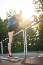 Woman stretches leg up on railings surrounded by trees in sunlight, middle distance runner