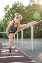 Woman stretches leg on high railing in sunshine on track, middle distance runner Katharina Jaiser,