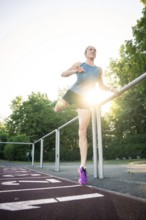 Woman stretching on a track in sunlight, holding onto the railing, surrounded by trees, middle