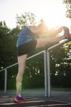 Woman stretches leg over railings surrounded by trees and sunlight, middle distance runner