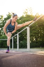 Woman stretches leg on railings in sunshine surrounded by trees near park, middle distance runner