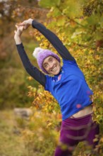Woman doing stretching exercises in autumn forest, smiling happily surrounded by colorful leaves,