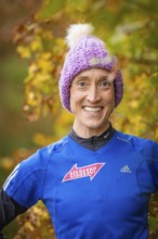 Woman wearing purple hat and blue sweater smiling in autumn surroundings, middle distance runner