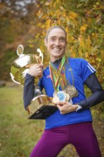 Radiant woman holding a big trophy and many medals in an autumn atmosphere, middle distance runner