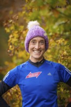 Person wearing blue clothes and purple cap smiling in autumn surroundings, middle distance runner