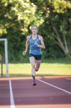Person running on a red track surrounded by green trees and bright sunlight, middle distance runner