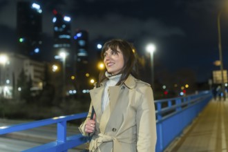 Woman wearing a trench coat and warm sweater standing on a bridge, looking at the illuminated city