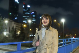 Young woman in a trench coat smiling as she walks across a city bridge at night, skyline lights and
