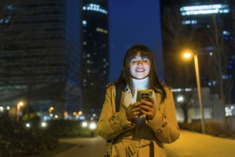 Young woman standing on an urban street at night, looking at her smartphone and smiling, with