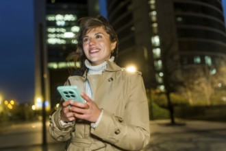 Woman smiling, holding a smartphone, and looking away in an illuminated urban environment with