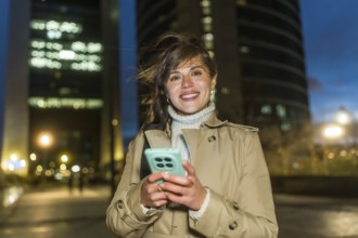 Young woman smiling, holding her smartphone, and looking at the camera outdoors in a modern city