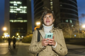 Woman standing outdoors at night, using a smartphone for communication and networking, dressed in a