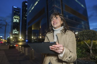 Woman stands at twilight in a glowing downtown, smiling while using a tablet, blurred skyscrapers