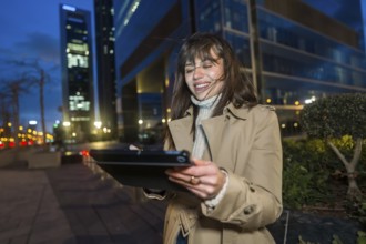 Young professional woman in a trench coat standing in a lit downtown at night, smiling while using