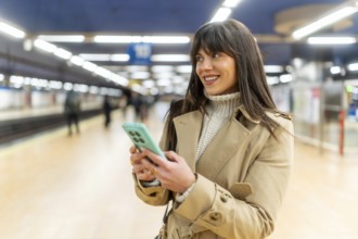 Woman standing on a train station platform, smiling and looking away while sending messages or