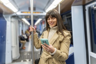 Young woman standing in a subway car, smiling at her smartphone screen during the commute, browsing