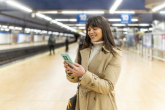Young woman standing on a subway platform and smiling while checking her mobile phone, actively
