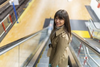 Woman in a trench coat smiling confidently while riding down an escalator in a modern subway