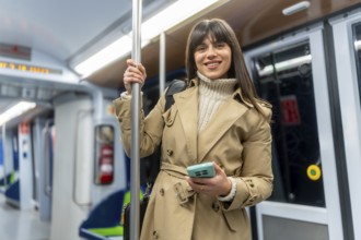 Young smiling woman in a trench coat stands in a subway car, holding her smartphone while commuting