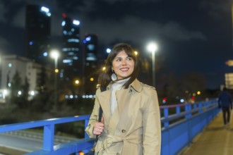 Woman in trench coat on an urban bridge at night, smiling and looking away as illuminated