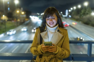 Young woman smiling and engaging with a mobile phone, standing on an urban bridge overlooking a