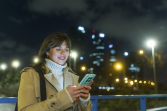 Young woman smiling as she types on her smartphone on a city street at night, illuminated by bokeh