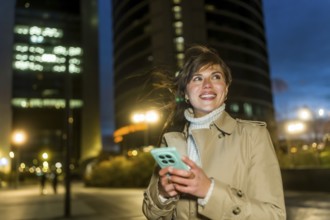 Young woman smiling, looking up while navigating her smartphone, dressed in a trench coat and