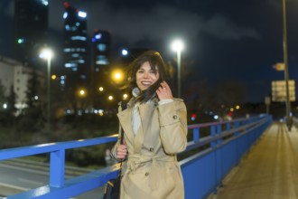 Young woman in a trench coat smiling, touching her hair, and looking at the camera while standing