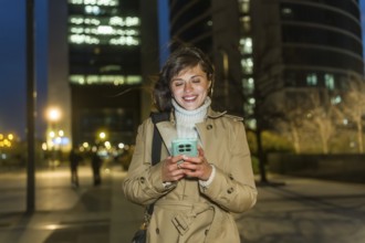 Young woman in a trench coat smiling, walking through a city at night, connecting with her