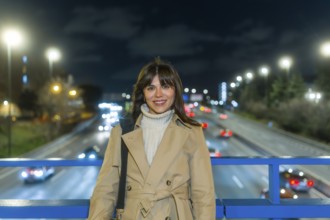 Woman smiling and standing on a bridge over a blurred highway with car lights creating bokeh,