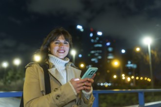 Woman standing on a city bridge at night, smiling and typing on her mobile phone, with illuminated