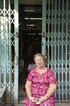 Bangkok, Thailand. March 18th 2025. A smiling older lady sitting outside a shop house in the