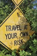 A weathered yellow road sign with the text 'Travel at Your Own Risk, 'flanked by leaves, Grand