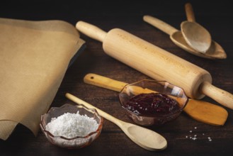 Rolling pin, cooking spoon and baking paper next to jam and sugar on a dark wooden table in a
