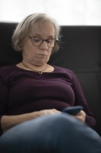 Elderly woman with glasses, gray hair, sitting on sofa and using smartphone, cell phone, Stuttgart,