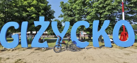 A bicycle stands in front of a large blue lettering of 'Gizycko' on a sandy beach, Masuria bicycle