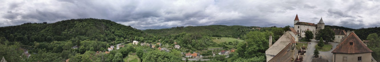 Panoramic view of a hilly landscape with a castle complex and thick greenery under cloudy sky,