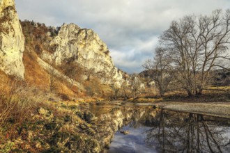 An autumn landscape with a river reflecting the surrounding rocks and trees, under a cloudy sky,