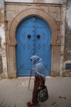 Tunis, Tunisia. 7th May 2024 An old woman wearing a blue shawl walks past a traditional blue door
