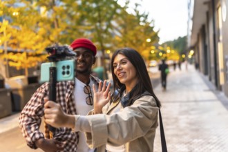 Diverse couple vlogging together on a city sidewalk, waving to camera held on a selfie stick,