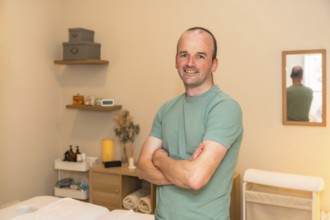 Professional man massage therapist smiling confidently, standing with crossed arms in a therapy