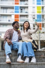 Happy diverse young couple sitting on concrete steps, smiling and taking a cheerful selfie together