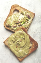 Two slices of toasted bread, topped with pistachio paste and sprinkled with pistachios, spread on a