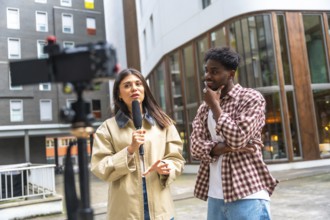 Female journalist holding a microphone and gesturing while interviewing a young man on a city