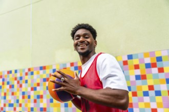 Young man smiling and holding a vibrant basketball in sportswear, standing against a colorful tiled